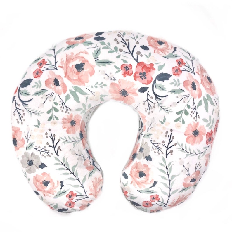 May include: A white nursing pillow with a floral print featuring pink and green flowers. The pillow is shaped like a crescent moon and is designed to support a baby during breastfeeding or bottle feeding.