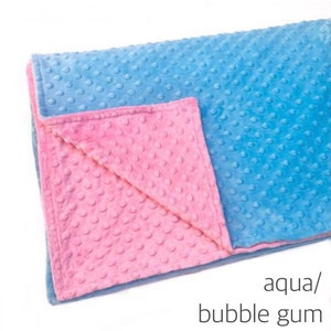 May include: A soft, textured blanket featuring a dimpled minky surface. One side is a bright aqua blue, while the folded corner reveals a contrasting bubble gum pink underside. The text aqua/bubble gum is printed in the bottom right corner.