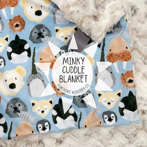 Puede incluir: Una manta para beb&eacute; con un fondo azul claro con caras de animales marinos de dibujos animados, incluyendo focas, zorros y ballenas. La manta tiene un borde suave y afelpado de color crema y el texto "Minky Cuddle Blanket".
