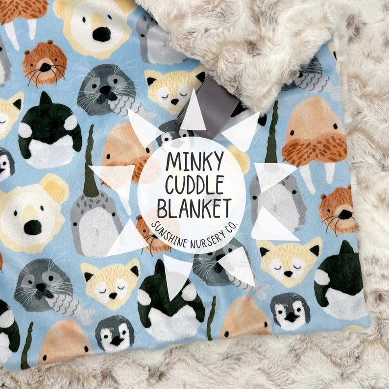 Puede incluir: Una manta para beb&eacute; con un fondo azul claro con caras de animales marinos de dibujos animados, incluyendo focas, zorros y ballenas. La manta tiene un borde suave y afelpado de color crema y el texto "Minky Cuddle Blanket".