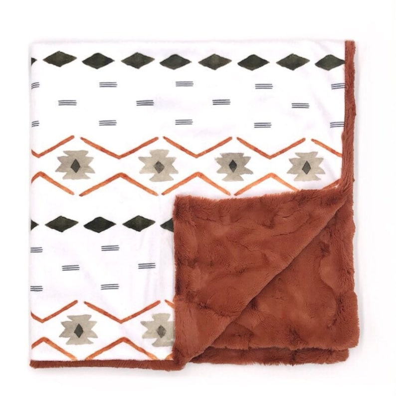 May include: A soft, plush blanket with a white background and a geometric pattern in shades of orange, green, and gray. The reverse side of the blanket is a solid rust color.