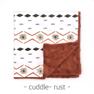 May include: A white and rust colored blanket with a geometric pattern. One side of the blanket is a soft, plush fabric, while the other side features a printed design. The blanket is labeled "cuddle - rust - "