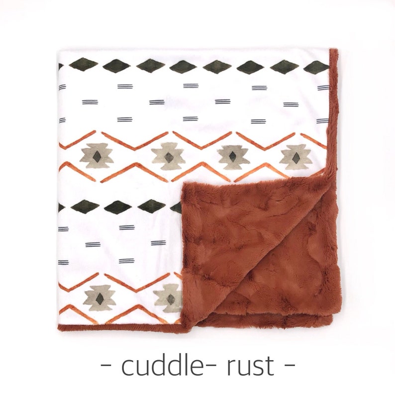 May include: A white and rust colored blanket with a geometric pattern. One side of the blanket is a soft, plush fabric, while the other side features a printed design. The blanket is labeled "cuddle - rust - "
