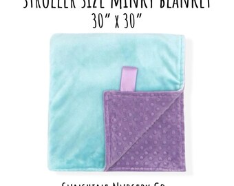 May include: A square minky blanket measuring 30 inches by 30 inches, featuring a smooth light blue side and a textured purple side with raised dots. A purple fabric tag is attached to the edge. Text reads: STROLLER SIZE MINKY BLANKET 30" x 30" SUNSHINE NURSERY CO.