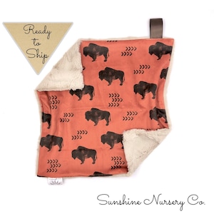 May include: A soft, plush baby blanket with a brown buffalo print on a terracotta background. The blanket has a white faux fur backing and a brown leather loop for hanging.