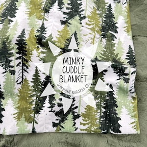 May include: A minky cuddle blanket with a forest pattern in shades of green, white, and black. The blanket features a repeating design of pine trees. The text "Minky Cuddle Blanket" and "Sunshine Nursery Co." are printed on the blanket.