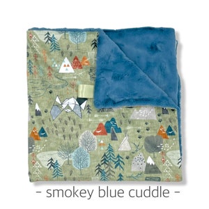 May include: A soft, double-sided blanket with a forest-themed pattern. The top layer features a green background with trees, mountains, and teepees. The bottom layer is a smoky blue plush material. A satin ribbon is attached. The text "- smokey blue cuddle -" is at the bottom.