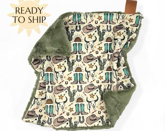 May include: A baby blanket with a cream-colored background featuring a repeating pattern of cowboy boots, hats, cacti, and bull skulls in brown, teal, and gold. The blanket has a soft, olive green plush border and a brown leather tag.