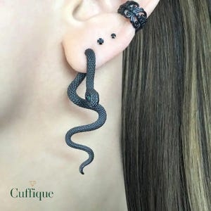May include: A dark grey snake earring with a green eye is shown. The earring is worn in a stretched earlobe. A black ear cuff and two small black studs are also visible on the ear. The earring is textured and has a curved design.