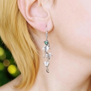 Può includere: Orecchini pendenti color argento con un design a forma di foglia. Ogni orecchino ha una piccola gemma verde e tre ciondoli a forma di foglia dettagliati. Gli orecchini sono mostrati su uno sfondo sfocato.
