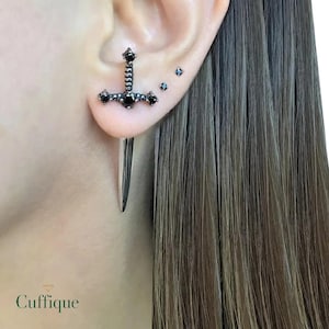 Pode incluir: Um brinco preto em forma de espada com pedras preciosas pretas. O brinco é usado na orelha, com uma longa lâmina pontiaguda pendurada abaixo do lóbulo. O brinco tem um design decorativo em forma de cruz com pedras preciosas pretas. A palavra "Cuffique" está na parte inferior.
