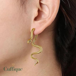 Peut inclure: Boucles d'oreilles serpent dorées. Les boucles d'oreilles s'enroulent autour de l'oreille, avec la tête du serpent en haut et le corps qui descend. Elles ont une surface texturée et l'œil du serpent est une petite pierre sombre. Le mot "Cuffique" est en bas.