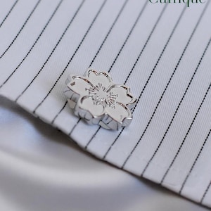 Könnte beinhalten: Ein silberfarbener Manschettenknopf in Blütenform liegt auf einem weißen Hemd mit schwarzen Nadelstreifen. Der Manschettenknopf hat detaillierte Blütenblätter und eine strukturierte Mitte. Das Wort "Cuffique" ist in grüner Schrift oben rechts gedruckt.