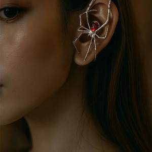 Pode incluir: Um brinco de orelha prateado em forma de aranha, com uma pedra vermelha no centro do corpo. O brinco é usado na orelha. A imagem é um close-up que destaca os detalhes da joia.