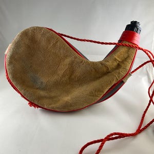 May include: A curved canteen featuring a tan suede exterior with red trim and a black plastic spout. A red braided rope is attached to the neck of the canteen, serving as a carrying strap. The overall shape is crescent-like with a textured surface.
