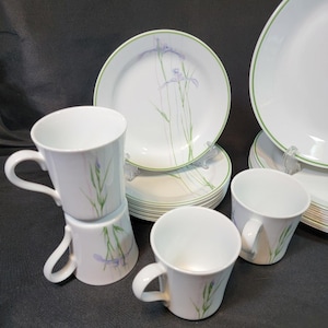 May include: A white dinnerware set featuring plates and mugs. The plates have a green border and a floral design with purple and green accents. The mugs have a matching floral pattern. The set includes multiple plates and mugs.
