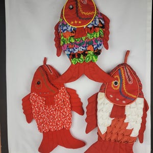 May include: Three decorative fish-shaped kitchen pot holders. The pot holders are primarily red, with colorful fabric accents and embroidered details. Each fish has a different pattern and design, adding a unique touch to the kitchen decor.