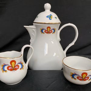 Könnte beinhalten: Ein weißes Porzellan-Kaffeeservice mit einem Blumenmuster in Rot, Gelb, Blau und Gold. Das Set umfasst eine Kaffeekanne mit Deckel, einen Sahnekännchen und eine kleine Schale. Die Ränder sind goldfarben.