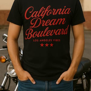 Può includere: T-shirt nero con la scritta "California Dream Boulevard" in rosso. Sotto il testo ci sono le parole "Los Angeles Vibes" e tre stelle rosse. La persona indossa jeans blu e si trova davanti a una moto.