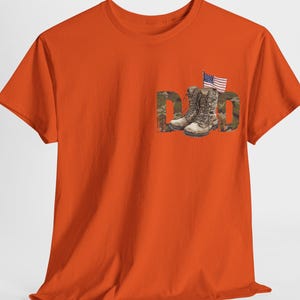 Può includere: T-shirt arancione con la scritta "DAD" in un design mimetico, accompagnata da un paio di anfibi e una piccola bandiera americana. La maglietta è realizzata in un materiale morbido e ha maniche corte.