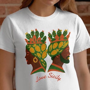 May include: White t-shirt featuring a colorful graphic of two side profiles wearing crowns with cactus and leaf designs. The text "Love Sicily" is printed in red cursive below the figures. The shirt is made of a soft material.