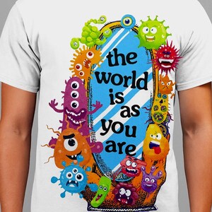 Può includere: T-shirt bianca con un colorato design a cartoni animati. Il design include uno specchio blu con la scritta "il mondo è come sei tu" circondato da vari mostri dei cartoni animati dai colori vivaci.