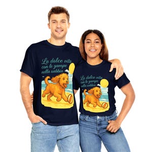 Può includere: T-shirt blu navy con grafica di un cane dorato che corre sulla spiaggia con una palla. Il testo "La dolce vita con le zampe nella sabbia" è sopra il cane. Le magliette sono indossate da un uomo e una donna.
