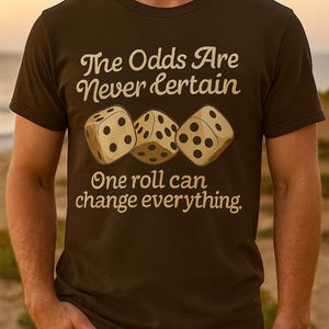 以下が含まれることがあります： 「The Odds Are Never Certain」と「One roll can change everything」の文字が入った茶色のTシャツ。最初のテキストの下には3つのサイコロが描かれています。サイコロはベージュ色で黒い点があります。