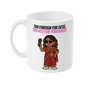 Könnte beinhalten: Weißer Keramikbecher mit einer Cartoon-Illustration einer Frau in einem Sari, die einen Spiegel hält. Der Becher trägt den Text "TOO FOREIGN FOR DESIS, TOO DESI FOR FOREIGNERS" in Rosa und Schwarz.