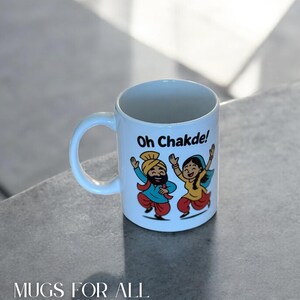 May include: White ceramic mug with a curved handle. The mug features a colorful illustration of two dancing figures in traditional attire, with the text "Oh Chakde!" and "Mugs for All Occasions".