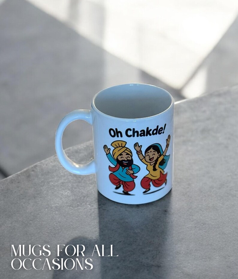 May include: White ceramic mug with a curved handle. The mug features a colorful illustration of two dancing figures in traditional attire, with the text "Oh Chakde!" and "Mugs for All Occasions".