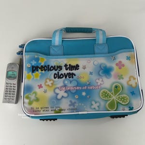 May include: A light blue and white laptop bag with a handle, featuring a floral design and the text "precious time clover". A silver cell phone is next to the bag. The bag has a clear front panel with flower and star graphics.
