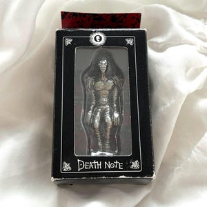 May include: A silver Death Note figurine in a black box. The box has a clear window showing the figure. The figure has long hair and is highly detailed. The box is labelled "Death Note" in white text.