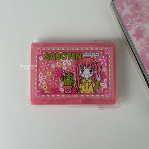 Puede incluir: Un estuche rectangular rosa con una ilustración de dibujos animados. El estuche presenta la palabra "SABOTEN" y un dibujo de una niña con pelo rosa y un cactus. El estuche está decorado con flores y corazones.