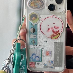 May include: A clear smartphone case decorated with various stickers, including a large pink character eating noodles, a small green creature, and several rectangular paper inserts. A teal lanyard with a star-shaped charm is attached to the side.
