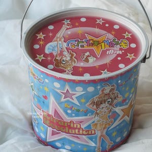 May include: A cylindrical metal bucket with a handle, featuring a pink lid with a cartoon character and stars. The blue sides display the text Kirarin Revolution, large stars, white polka dots, and a cartoon character in a yellow dress.