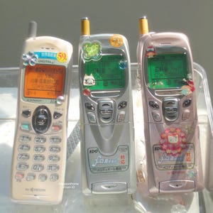 Puede incluir: Tres teléfonos móviles vintage en crema, plateado y rosa. Cada teléfono tiene una pantalla que muestra números y texto japonés. Los teléfonos están decorados con pegatinas y abalorios. El teléfono crema tiene el número 59.