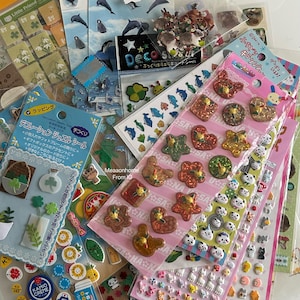 Puede incluir: Una colección de hojas de pegatinas coloridas con varios diseños, incluyendo animales, plantas y formas decorativas. Algunas pegatinas tienen purpurina y otras texto japonés. Las pegatinas son de varios tamaños y formas, perfectas para manualidades.