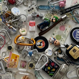 Puede incluir: Una colección de llaveros variados con diversos diseños, incluyendo personajes de dibujos animados, objetos en miniatura y amuletos temáticos de viajes. Los llaveros están hechos de metal, plástico y otros materiales, con una variedad de colores y formas. Algunos llaveros tienen texto, incluyendo "HAWAII" y "COLOMBIA".