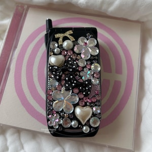 Puede incluir: Teléfono móvil negro adornado con pedrería transparente y rosa, perlas y un lazo. El teléfono presenta formas de flores y corazones. El teléfono está encima de una caja de CD con un diseño rosa y blanco.