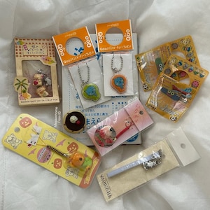 Puede incluir: Una colección de merchandising de personajes japoneses, que incluye llaveros, una correa y un charm con forma de postre. Los artículos presentan personajes de dibujos animados y están empaquetados en varios colores, como amarillo, naranja y rosa.