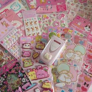 May include: A collection of pink and pastel stickers featuring various cartoon characters, including Hello Kitty, Bambi, and other cute designs. A pink flip phone is also present, along with Japanese text.