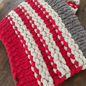 May include: A chunky knit blanket in red, gray, and off-white. The blanket features thick, braided stripes of red and off-white, with a solid gray section. The cozy, handmade blanket is folded on a wooden surface.