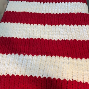 May include: A chunky knit blanket featuring alternating wide stripes of red and white. The blanket has a soft, textured appearance, with each stripe clearly defined. The color scheme is bold and eye-catching, suitable for a cozy home decor item.