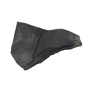 May include: A jagged, irregular piece of dark charcoal-colored stone with a matte, textured surface. The object features sharp, broken edges and a flat, slightly angled face, displaying natural variations in its dark gray and black coloration.