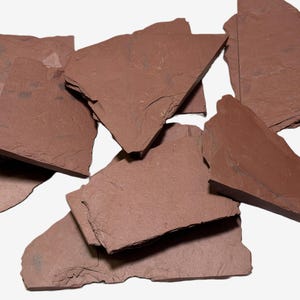 May include: A collection of irregularly shaped, reddish-brown slate pieces. The natural stone fragments vary in size and thickness, with rough, textured surfaces and jagged edges. The pieces are scattered against a white background.