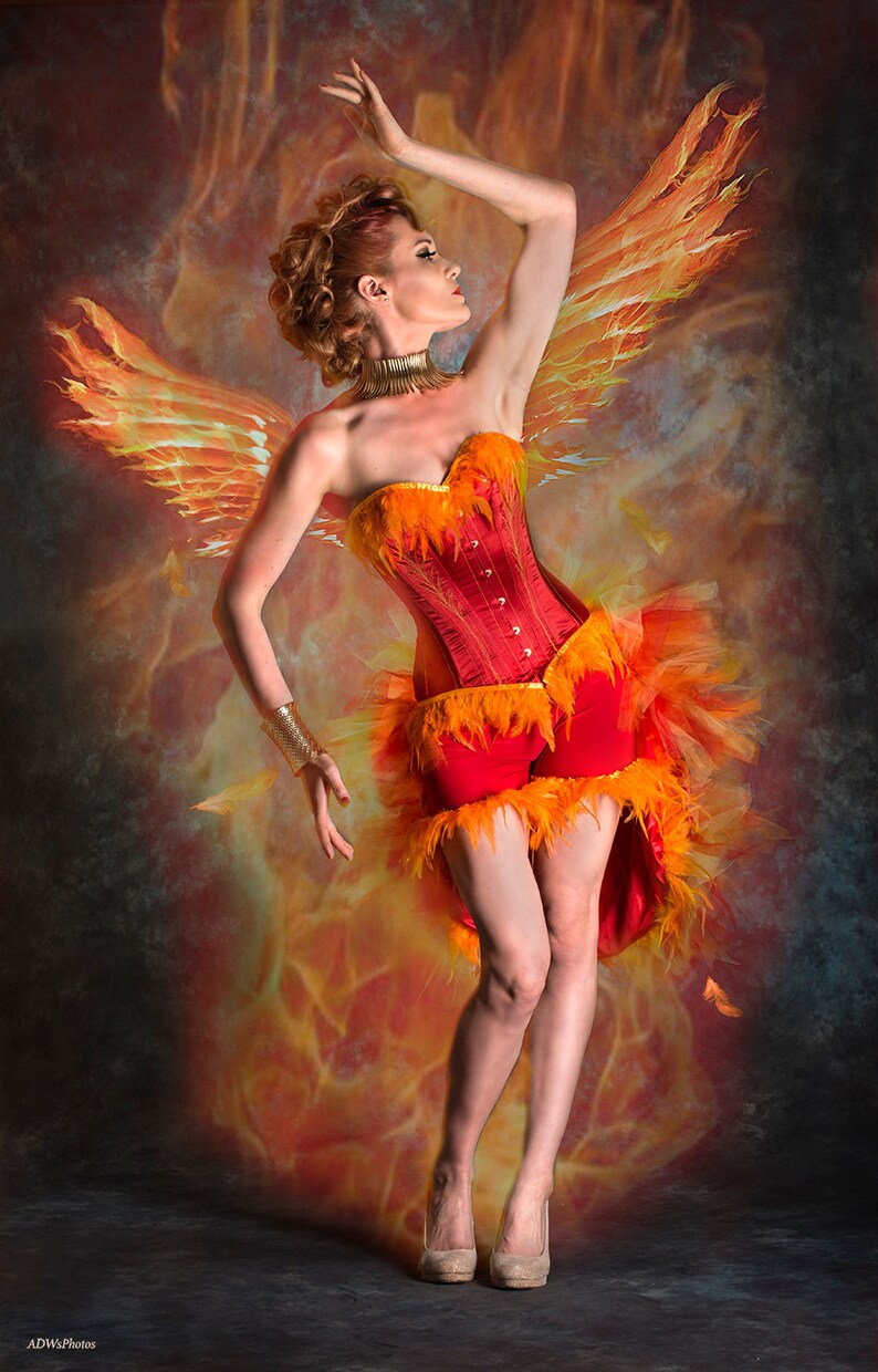 May include: A woman wearing a red and orange corset with feathers, standing in front of a fiery background. She is wearing a gold bracelet and has her hair styled in a bun.