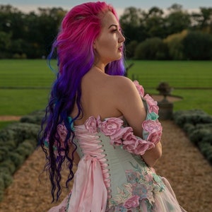 Puede incluir: Una mujer con cabello rosa y morado lleva un cors&eacute; verde claro y rosa con bordados florales y una cinta rosa. El cors&eacute; es parte de un vestido m&aacute;s grande con una falda de tul.