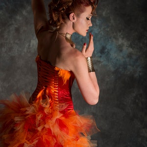 May include: A woman wearing a red and orange corset with a tulle skirt. She is wearing a gold necklace and bracelet. The background is a dark gray.