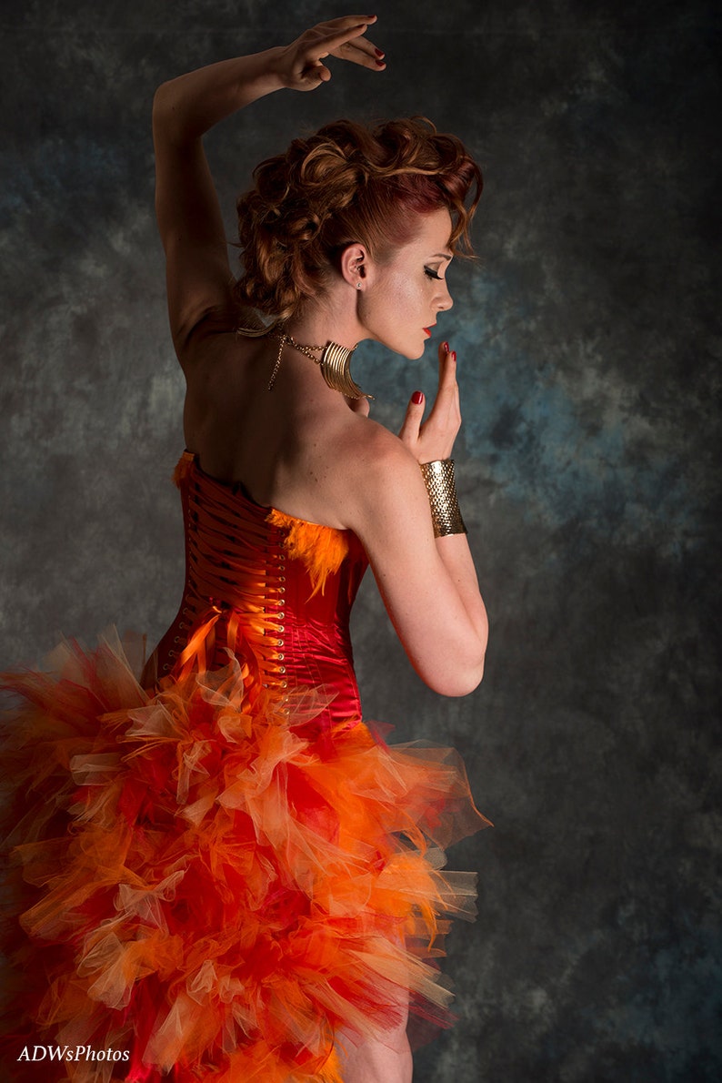 May include: A woman wearing a red and orange corset with a tulle skirt. She is wearing a gold necklace and bracelet. The background is a dark gray.